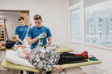 Fizioterapeits palīdz vecāka gadagājuma sievietei ar roku vingrinājumiem, kamēr viņa guļ uz izmeklējumu galda, bet cits terapeits strādā ar pacientu fonā. Izskatās, ka vide ir spilgta terapijas klīnika.