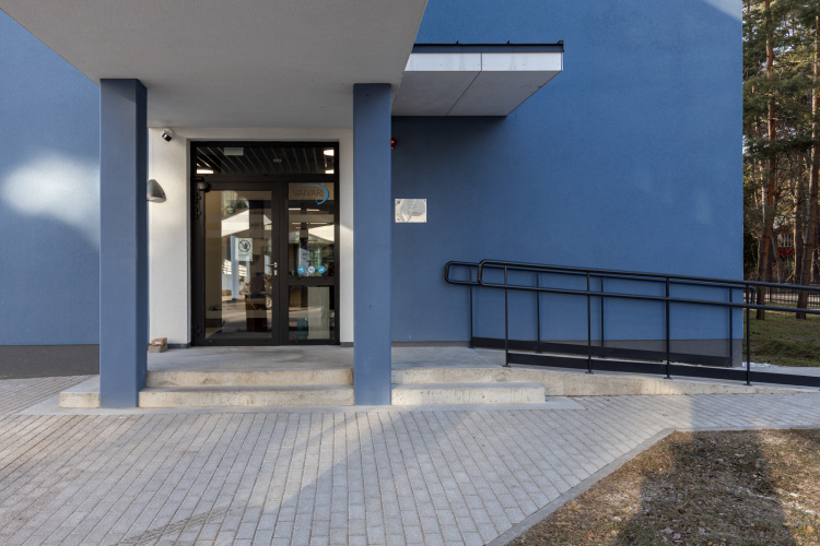 Mūsdienīga ēkas ieeja ar stikla durvīm, betona uzbrauktuve ar melnām margām ratiņkrēslu pieejamībai un līdzens celiņš. Ārējās sienas ir zilā krāsā, un fonā redzami koki.