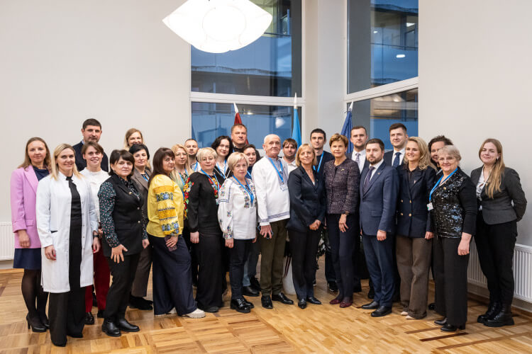 Cilvēku grupa stāv kopā iekštelpās un pozē fotogrāfijai. Daži valkā uzvalkus, citi valkā tradicionālus izšūtus apģērbus. Fonā ir karogi, un viņi smaida kamerā uz lielo logu fona.