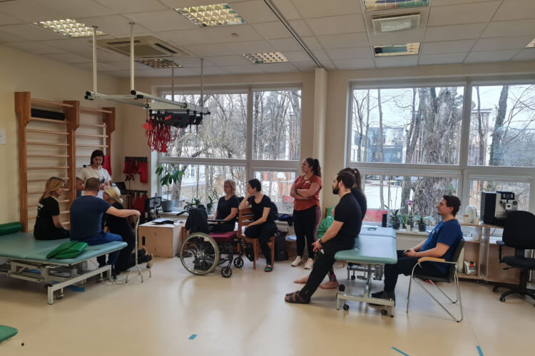 Cilvēku grupa, tostarp viens ratiņkrēslā, ir sapulcināts rehabilitācijas telpā ar vingrošanas galdiem un aprīkojumu. Šķiet, ka viņiem ir diskusija. Lielie logi atklāj kokus un ēku ārpusē.