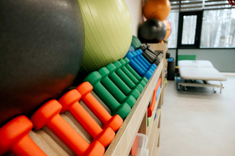 Krāšņu hanteļu un vingrošanas bumbu rinda ir glīti sakārtota plauktos gaišā telpā ar masāžas galdu pie loga fonā.