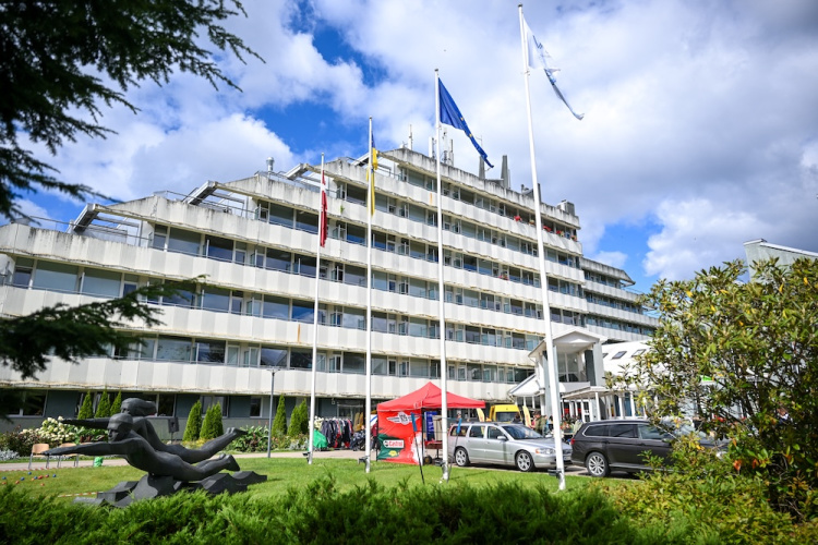 Mūsdienīga daudzstāvu ēka ar balkoniem, vairāki karoga masti priekšā, melna skulptūra zālienā un sarkana telts ar izstādītām drēbēm netālu, daļēji apmākušās debesis.