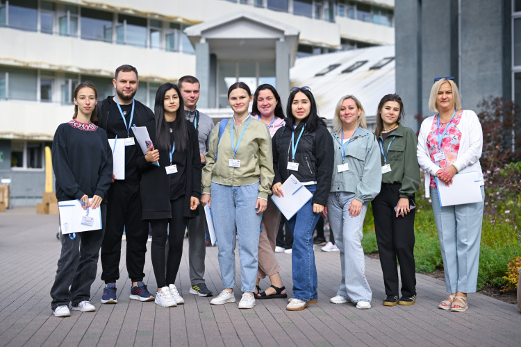 Desmit cilvēku grupa, kuriem rokās ir vārda uzlīmes un papīri, smaidot stāv uz bruģēta celiņa pie modernas ēkas.