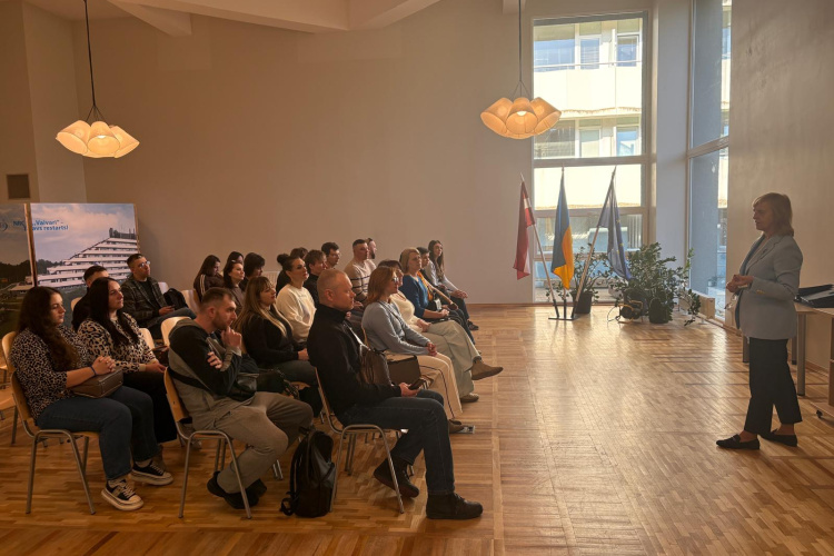 Sieviete gaiši zilā kostīmā stāv un uzrunā klausītājus, kas sēž rindās gaišā telpā ar lieliem logiem, koka grīdām un vairākiem karogiem un augiem fonā.