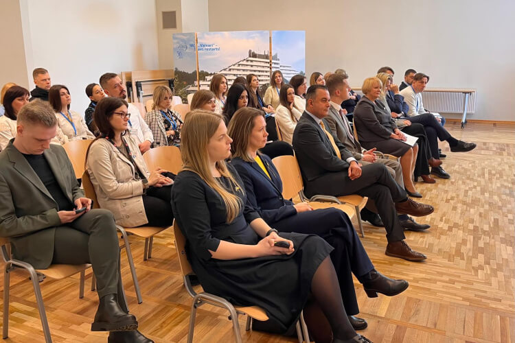 Cilvēku grupa, kas sēž rindās konferenču telpā, uzmanīgi klausās prezentāciju. Fonā redzams plakāts ar arhitektūras attēliem. Numurā ir koka grīda un baltas sienas.