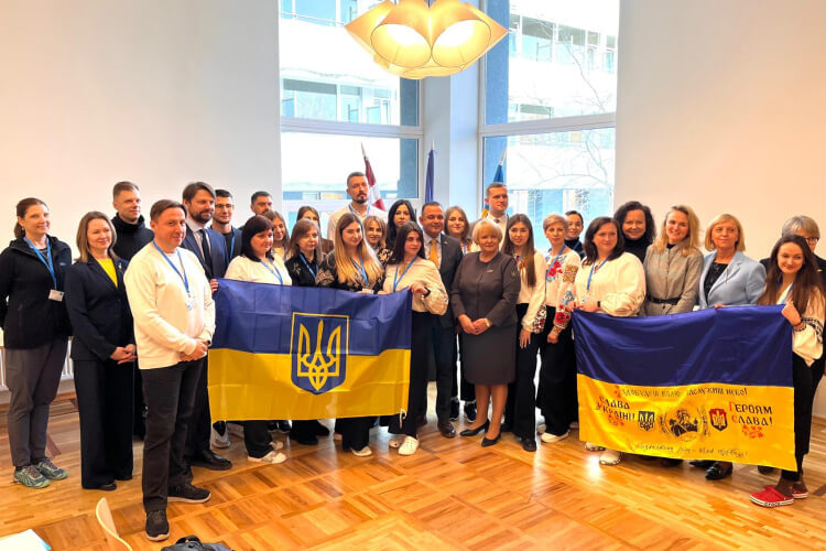 Cilvēku grupa iekštelpās pozē ar diviem Ukrainas karogiem. Tie stāv liela loga un modernas lustras priekšā. Daži cilvēki valkā tradicionālus izšūtus apģērbus. Istabā ir koka grīda.