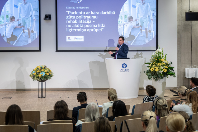 Vīrietis uzstājas pie tribīnes klīniskās konferences laikā Latvijas Universitātē. Aiz viņa redzams prezentācijas slaids latviešu valodā, un uz skatuves ir divi lieli ziedu pušķi.