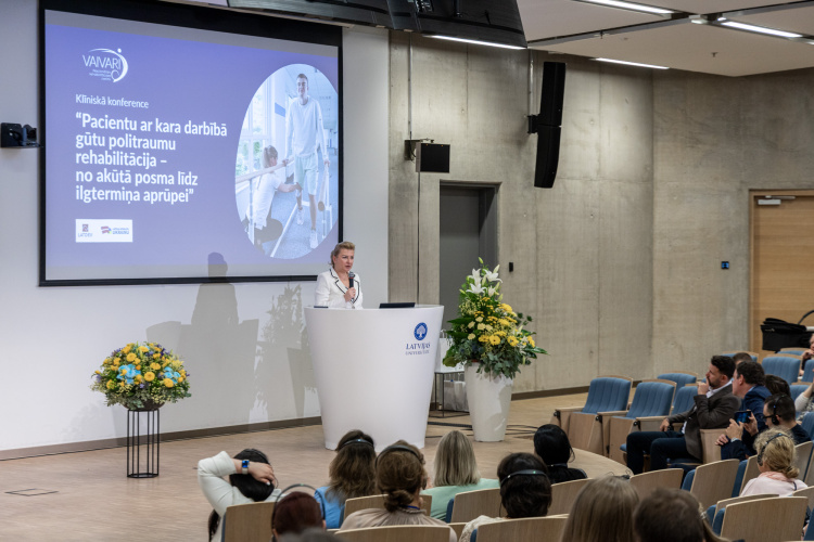 Klīniskajā konferencē sieviete uzstājas pie pjedestāla modernā lekciju zālē, aiz viņas sēž auditorija un uz liela ekrāna tiek rādīta prezentācija latviešu valodā.