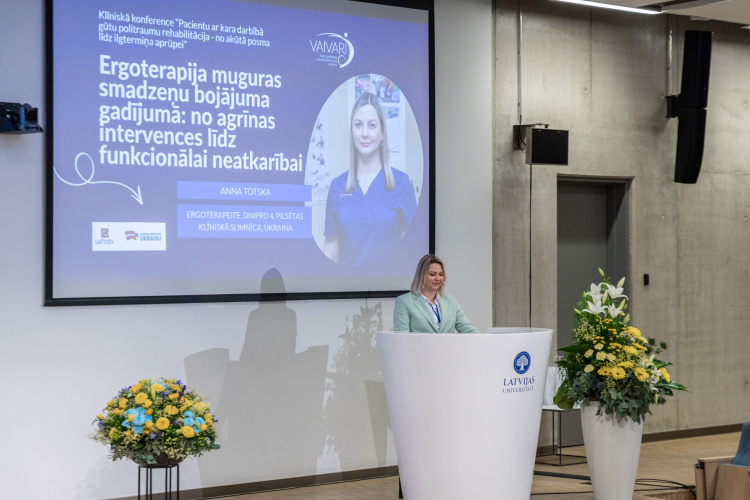 Sieviete stāv pie baltas tribīnes un uzstājas konferencē. Aiz viņas ir liels ekrāns, uz kura redzama prezentācijas slaida ar viņas fotogrāfiju un informāciju latviešu valodā. Skatuvi rotā ziedu kompozīcijas.