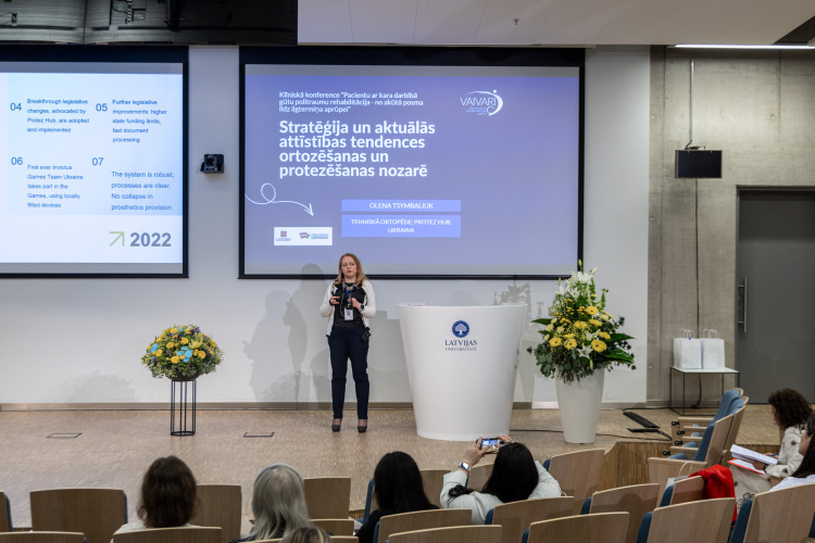 Uz skatuves stāv sieviete, kas uzstājas uz skatuves un uzstājas ar prezentāciju modernā konferenču zālē. Aiz viņas ir liels ekrāns ar tekstu latviešu valodā un ziedu kompozīcijām. Vairāki konferences dalībnieki sēž, vērsti pret runātāju.