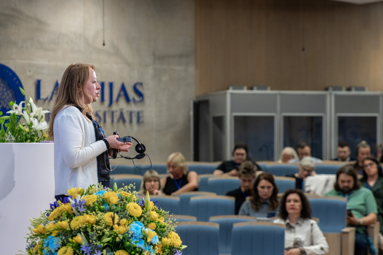 Sieviete stāv un runā pie pjedestāla ar ziediem, uzrunājot auditoriju, kas sēž zilos krēslos modernā lekciju zālē. Daži cilvēki auditorijā, šķiet, uzmanīgi klausās.