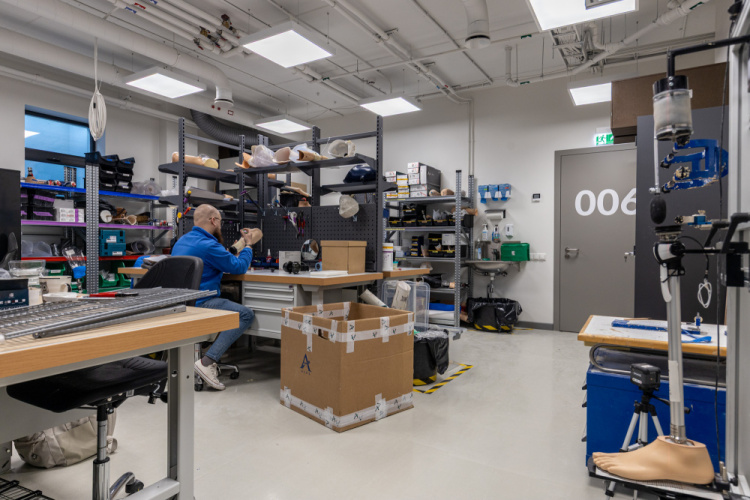 Cilvēks sēž pie darba vietas labi apgaismotā laboratorijā, kas pilna ar protēzēm, instrumentiem un aprīkojumu. Plauktos ir dažādi materiāli, un telpas centrā uz grīdas ir novietota liela kartona kaste.