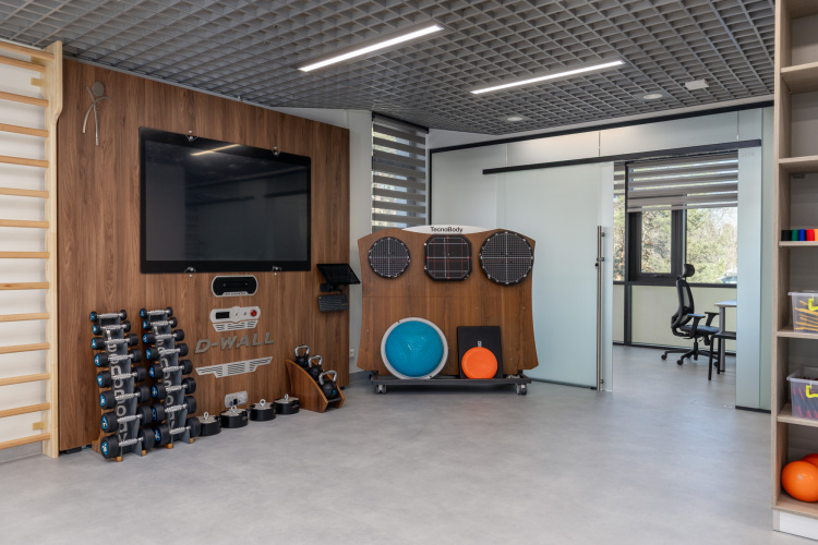 Moderns rehabilitācijas vai fitnesa kabinets ar trenažieriem, tostarp hantelēm, līdzsvara dēļiem, medicīniskām bumbām un digitālo treniņu sienu; pa stikla durvīm ir redzams kabinets.