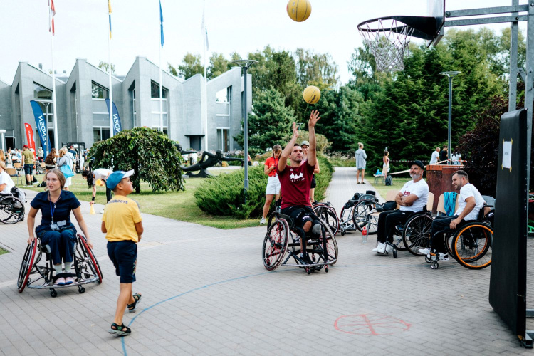Vairāki cilvēki ratiņkrēslos spēlē basketbolu brīvā dabā uz bruģēta laukuma. Spēlētājs sarkanā krekliņā met bumbu grozā, kamēr pārējie skatās. Blakus stāv bērns dzeltenā krekliņā. Fona plānā redzamas ēkas un koki.