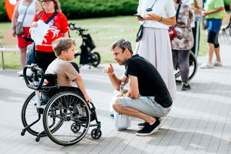 Vīrietis sēž tupus un sarunājas ar smaidošu zēnu ratiņkrēslā ārā, apjozts ar citiem cilvēkiem. Atmosfēra šķiet draudzīga un sabiedriska.