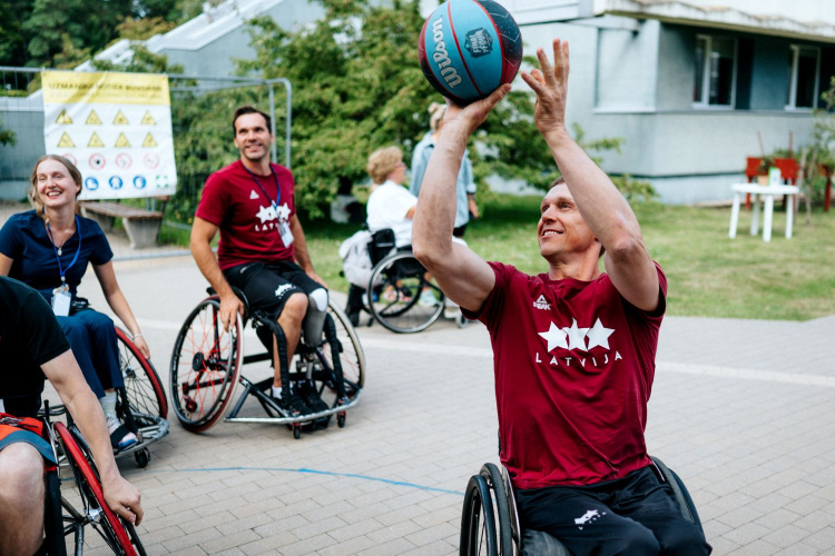 Četri cilvēki ratiņkrēslos spēlē basketbolu brīvā dabā. Vīrietis sarkanā Latvijas krekliņā met bumbu, kamēr pārējie skatās un smaida. Fona redzami koki un ēkas.