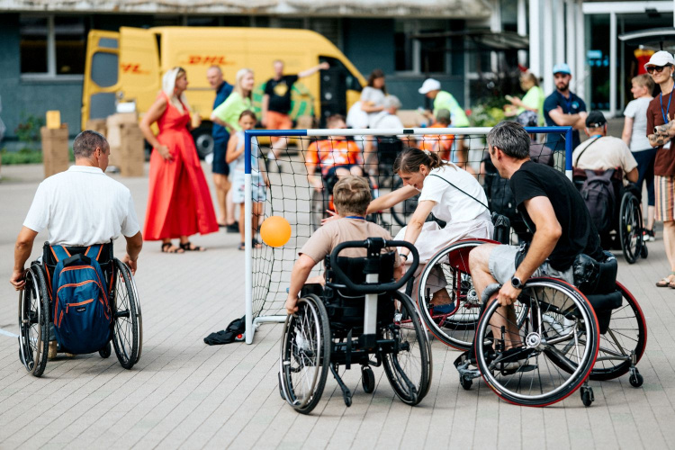 Cilvēki ratiņkrēslos ārā spēlē spēli ar nelieliem vārtiem, kamēr citi to vēro fonā. Redzams dzeltens mikroautobuss un cilvēki spilgtās drēbēs.