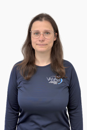 Sieviete ar gariem brūniem matiem un brillēm, tērpusies tumši zilā krekliņā ar „Vaivari“ logotipu, stāv priekšā vienkāršam baltam fonam, skatoties kamerā ar neitrālu sejas izteiksmi.