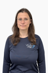 Sieviete ar gariem brūniem matiem, brilles un tumši zilu kreklu ar uzrakstu „VAIARI“ stāv uz vienkārša balta fona un ar neitrālu sejas izteiksmi skatās tieši kamerā.