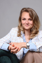 Sieviete gaišmatainiem matiem gaišā pledā, zilā kreklā un smilškrāsas biksēs sēž uz tumši zaļa dīvāna, smaida un noliek rokas sev priekšā. Fons ir vienkāršs un pelēks.