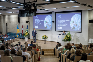 Uz skatuves konferencē auditorijā uzstājas vīrietis un sieviete. Klausītāji sēž, un uz diviem lieliem ekrāniem tiek rādīts viens un tas pats diapozitīvs ar vīrieti laboratorijas halātā, tekstu un vairāku valstu karogiem.