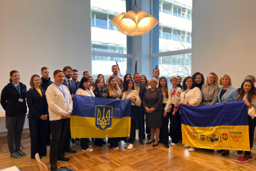 Cilvēku grupa stāv iekštelpās, turot rokās divus Ukrainas karogus. Viņi smaida, un daži ir ģērbušies tradicionālos izšūtos kreklos. Pie griestiem karājas moderns gaismas ķermenis, un lieli logi atklāj biroja ēku ārpusē.