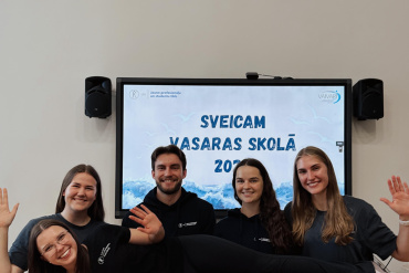 Pieci smaidoši cilvēki pozē telpā; trīs no viņiem priekšā horizontāli tur sievieti. Aiz viņiem redzams ekrāns, uz kura attēlots teksts latviešu valodā: „SVEICAM VASARAS SKOLĀ 2023”. Grupa izskatās laimīga un viesmīlīga.