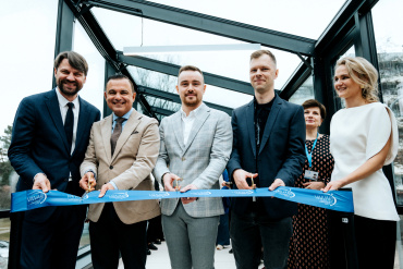 Seši cilvēki svinīgā tērpā stāv rindā un smaida, turot rokās un pārgriežot zilu lentīti svinīgā lentes griešanas ceremonijā. Daži no viņiem tur šķēres, kamēr citi tur lentīti.