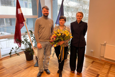 Trīs cilvēki stāv telpās pie lieliem logiem un karogiem. Sieviete centrā tur ziedu pušķi, bet divi cilvēki viņai blakus smaida. Ārā redzams sniegs.