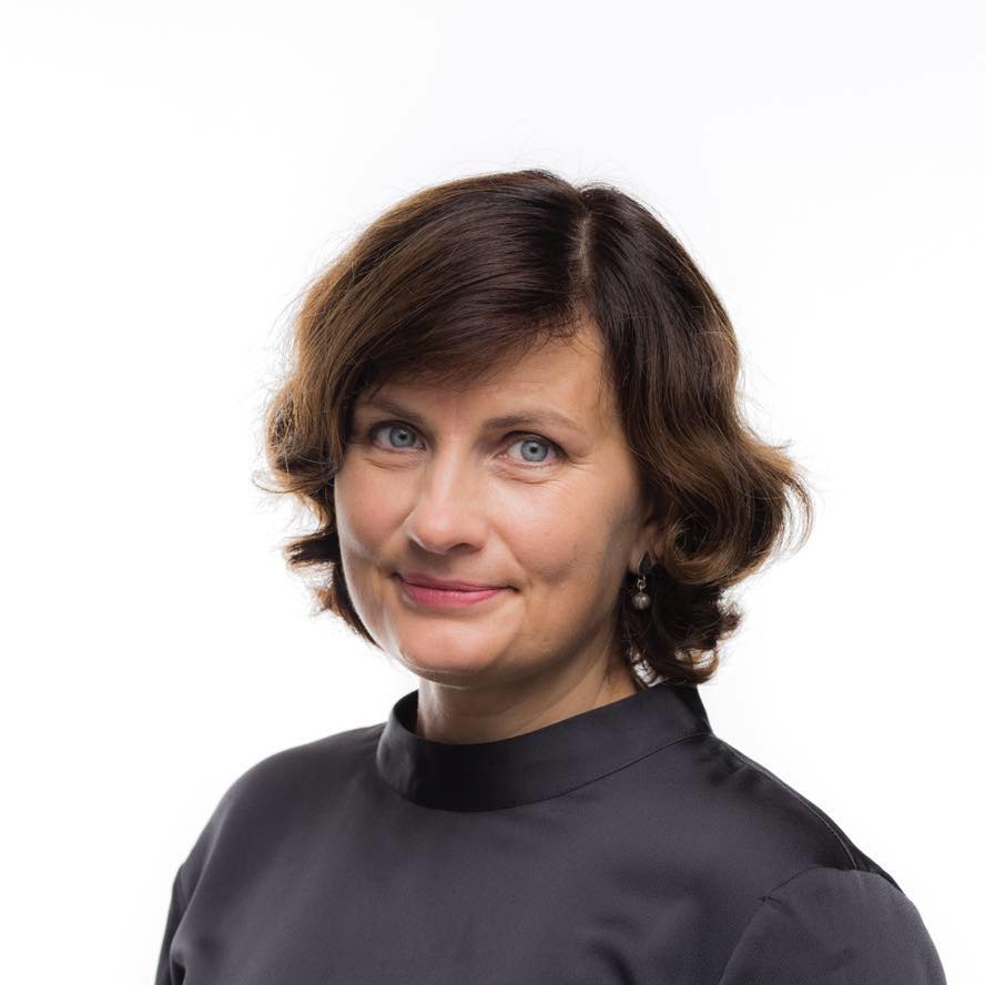 Sieviete ar īsiem, viļņainiem, brūniem matiem un zilām acīm nedaudz smaida. Viņa ir tērpusies melnā tērpā un mazos auskaros, fotografēta uz vienmuļa balta fona.